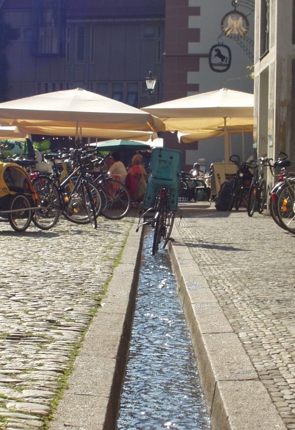 Freiburger Bächle in einer Kopfsteinpflasterstraße der Altstadt mit Fahrrädern und Straßencafés.