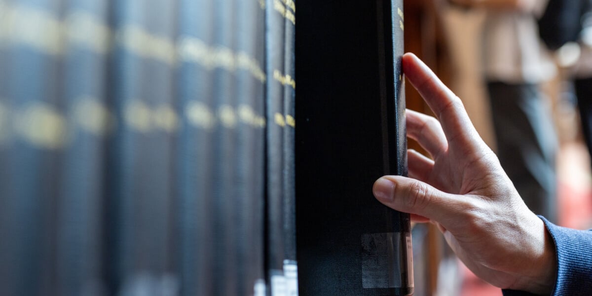 Eine Hand greift in einem schwach beleuchteten Buchladengang zwischen Bücherreihen in einem Bibliotheksregal nach einem dunklen Buch und erzeugt so eine ruhige, nachdenkliche Stimmung.