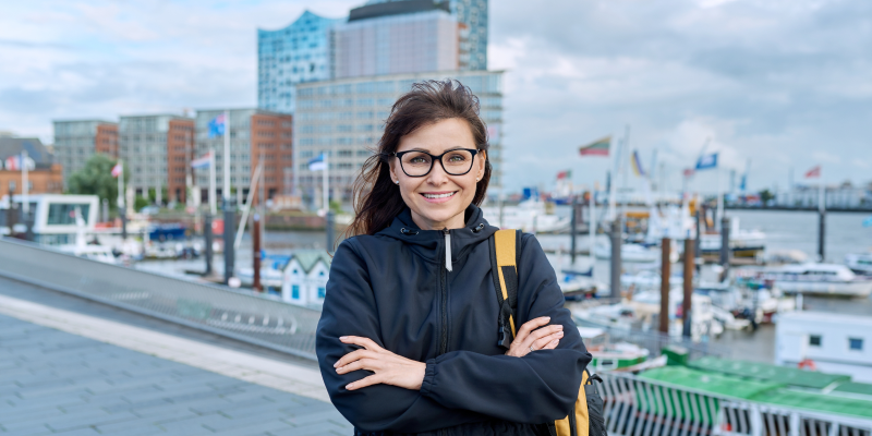 Lächelnde Frau mit Rucksack vor dem Hamburger Hafen und der Elbphilharmonie im Hintergrund.